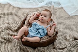 Baby in blue dress romper lying in wooden bowl on knit blanket during professional photoshoot.