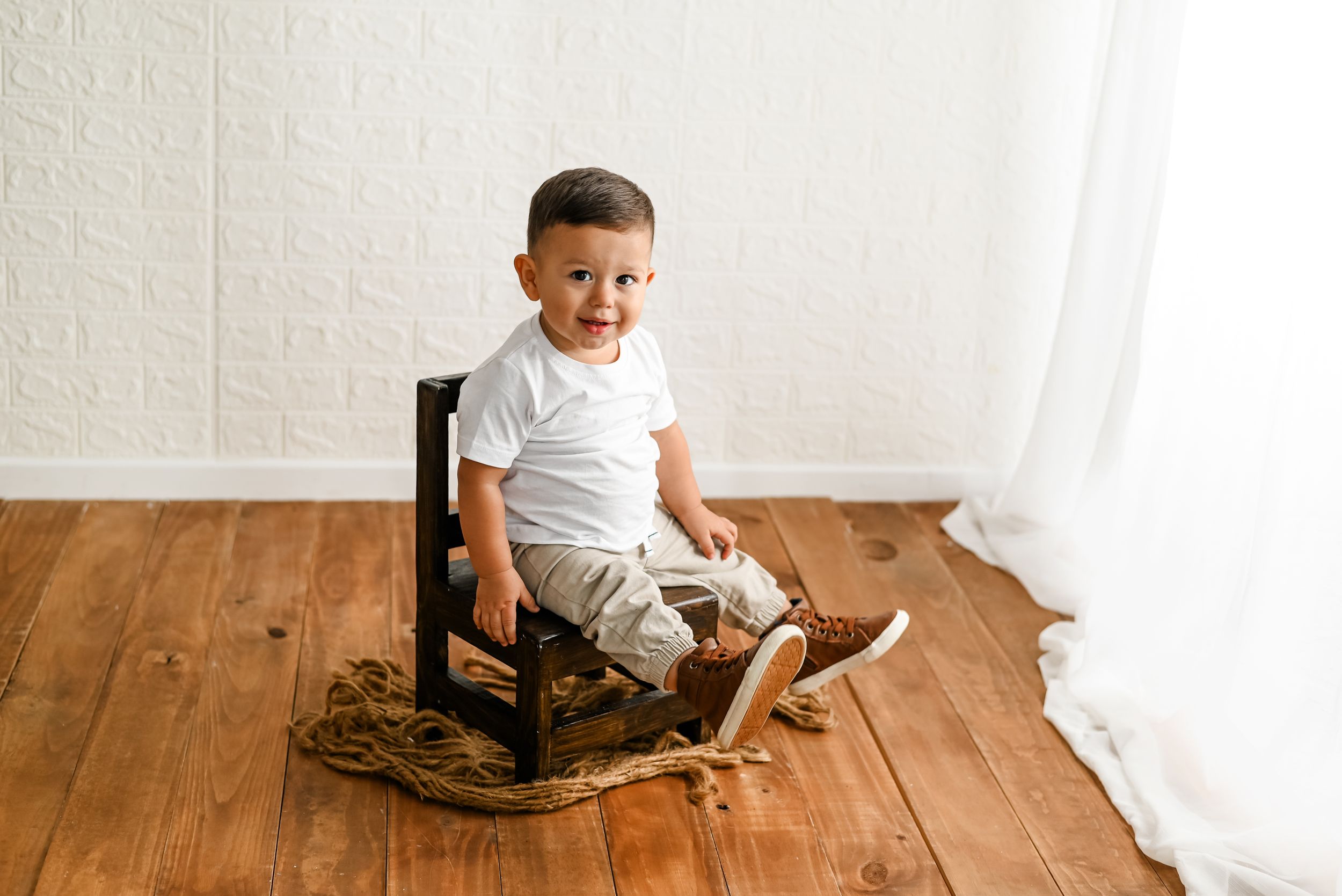 A small child sits in a dark wooden rocking chair on hardwood flooring against a white wall.