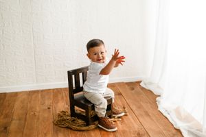 Small child in white clothing stands playfully on a wooden chair against white wall on hardwood floors.