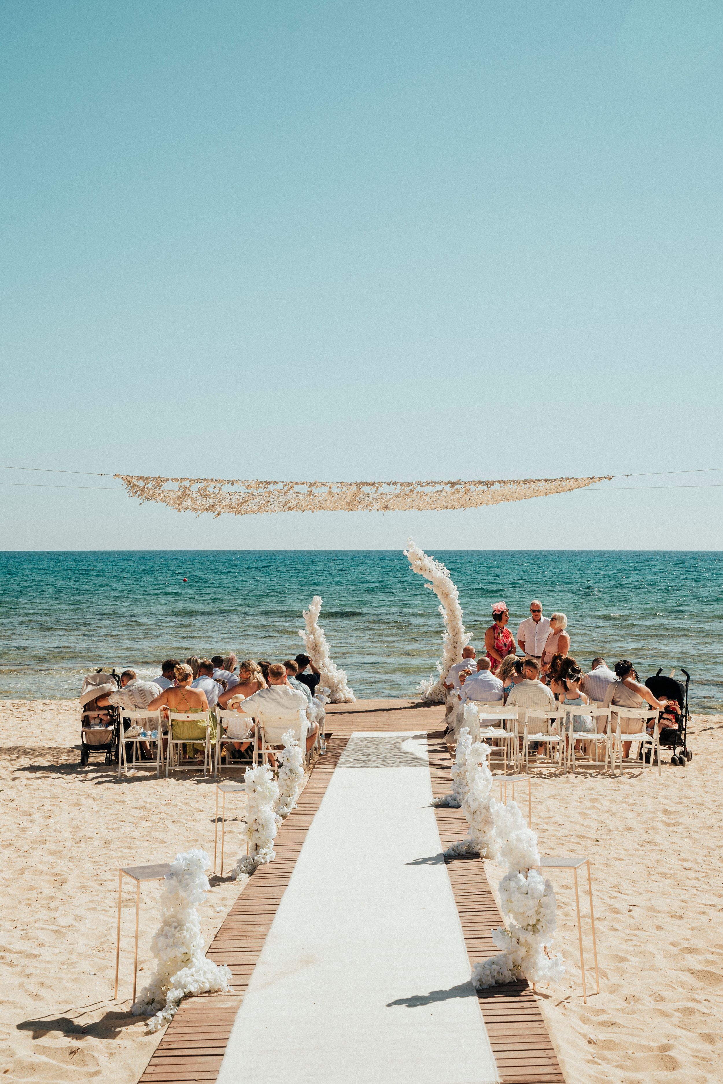 A romantic beach wedding ceremony setup with a white aisle runner and floral decorations along the ocean shore.