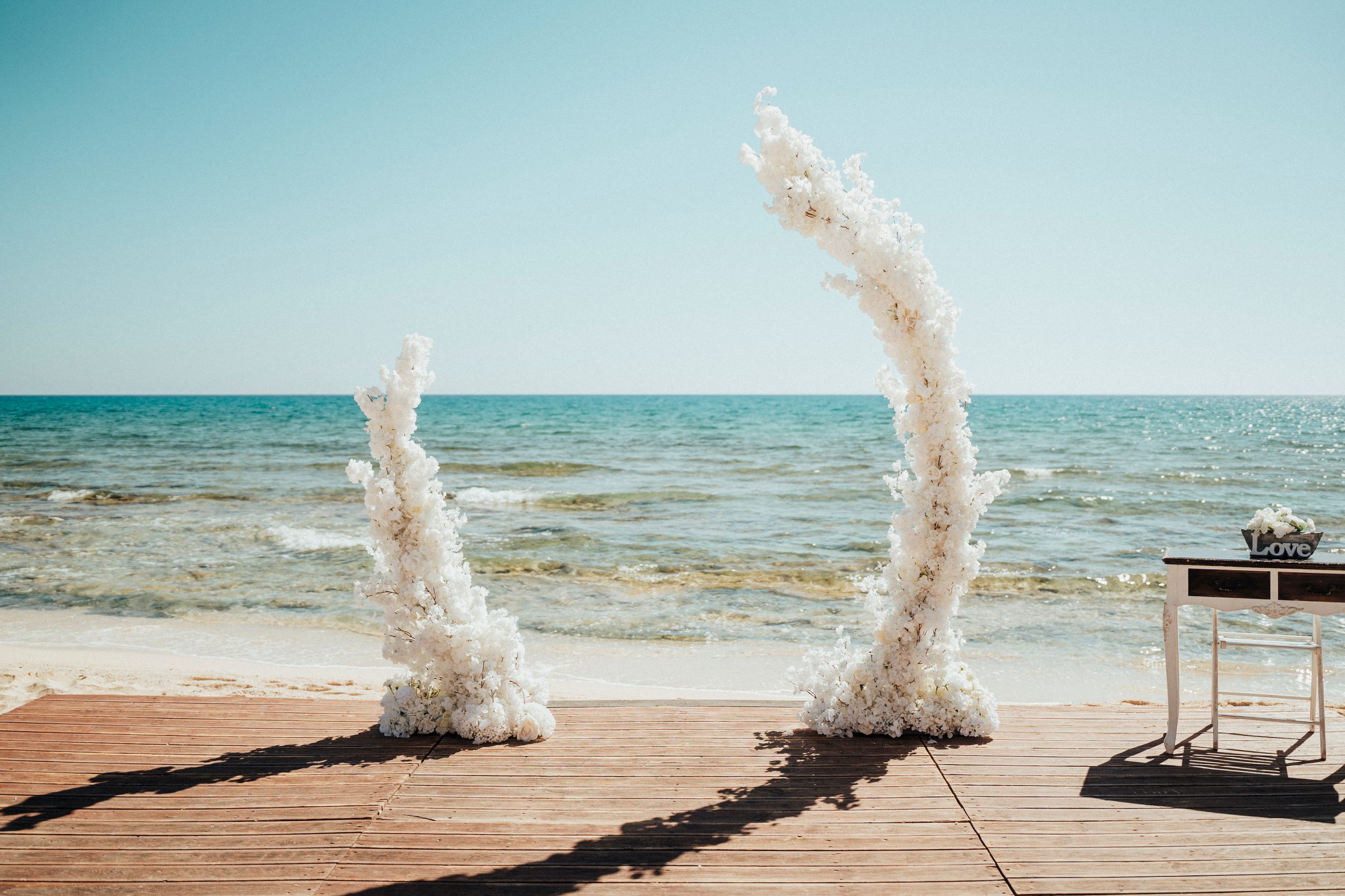 White pampas grass decorations frame a beachside ceremony setup with turquoise ocean waters in the background.