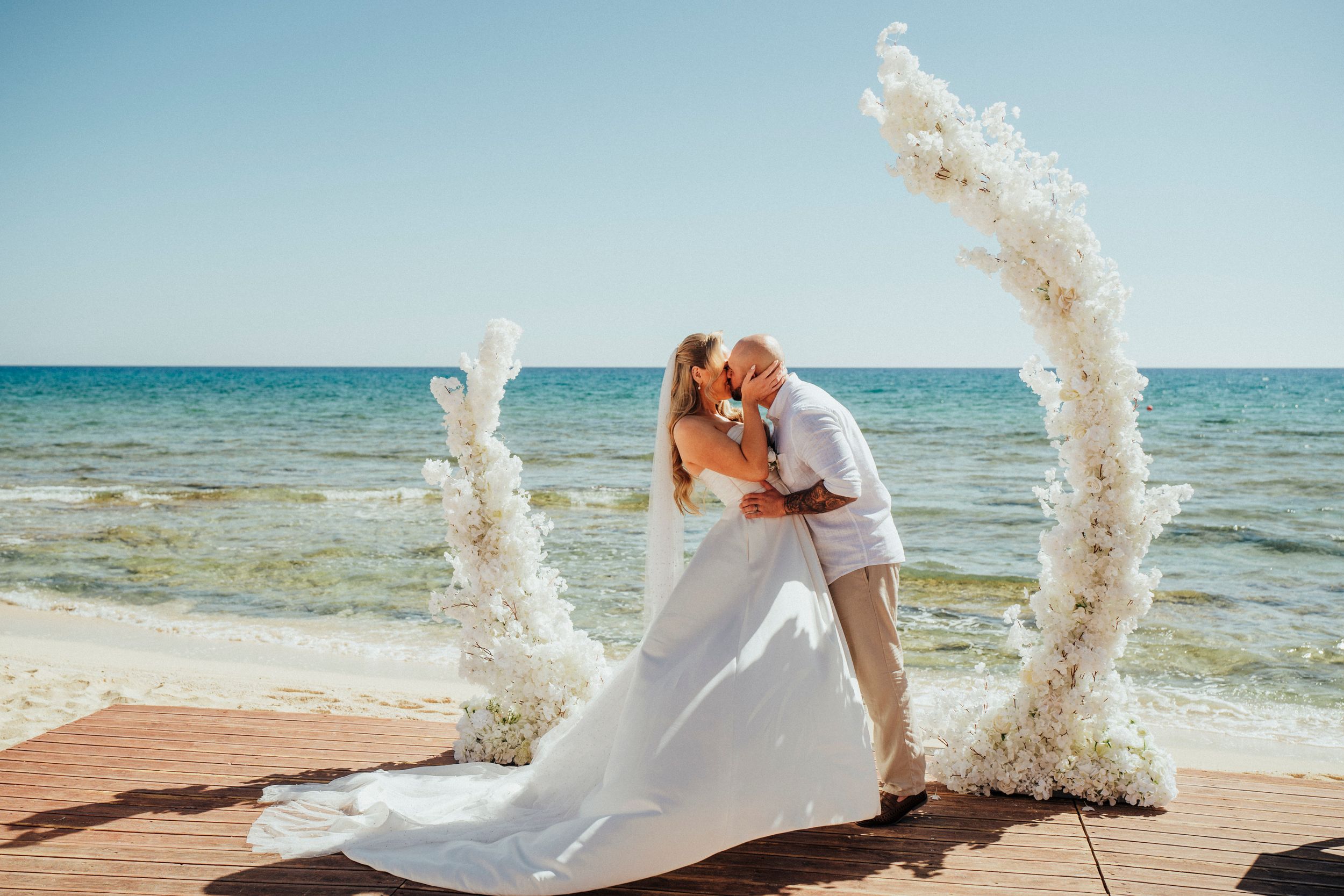 A couple shares a romantic kiss while splashing water creates dramatic arcs on a sunny beach by the blue ocean.