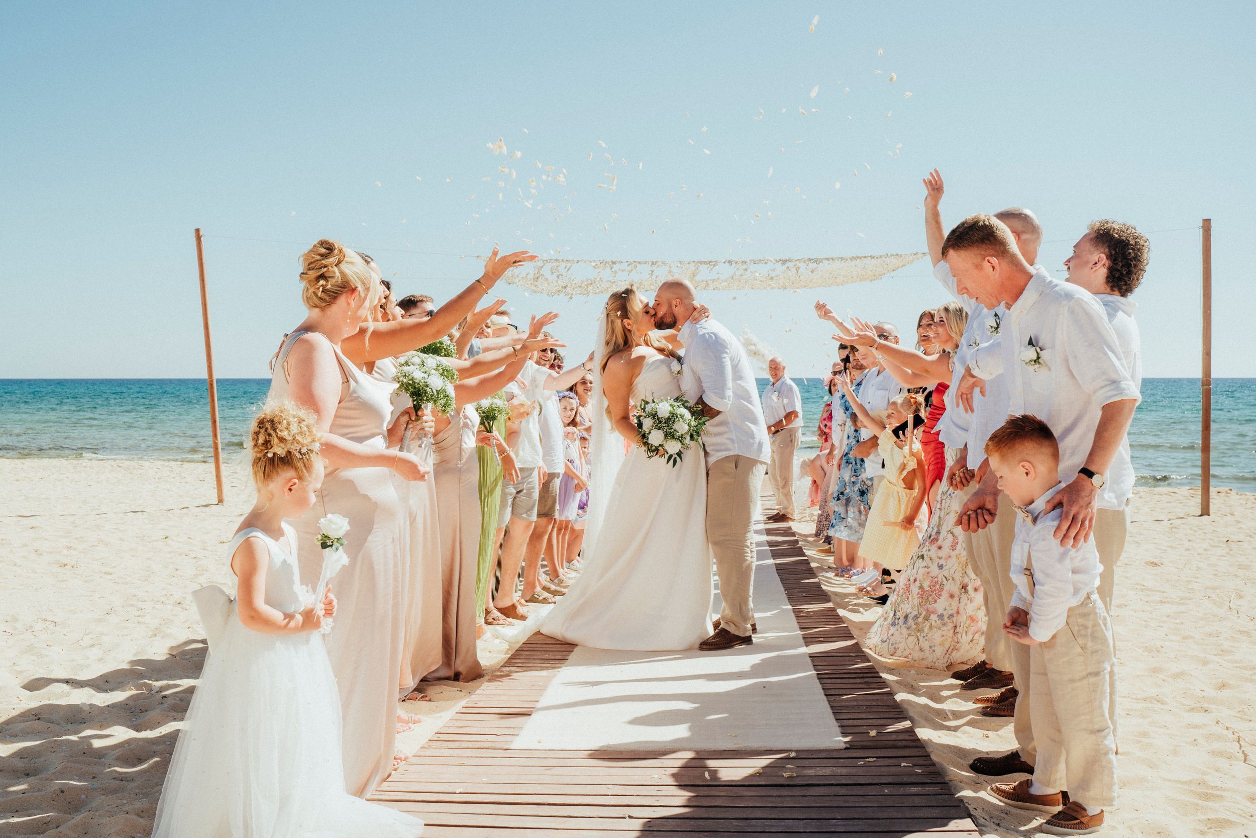Wedding celebration on a beach boardwalk with guests tossing confetti as a couple walks beneath.