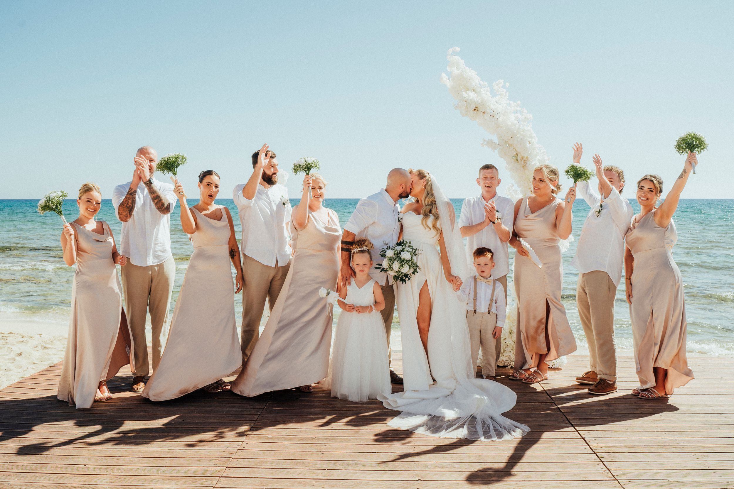 A joyful wedding party poses together on a wooden boardwalk by the ocean on a bright sunny day.