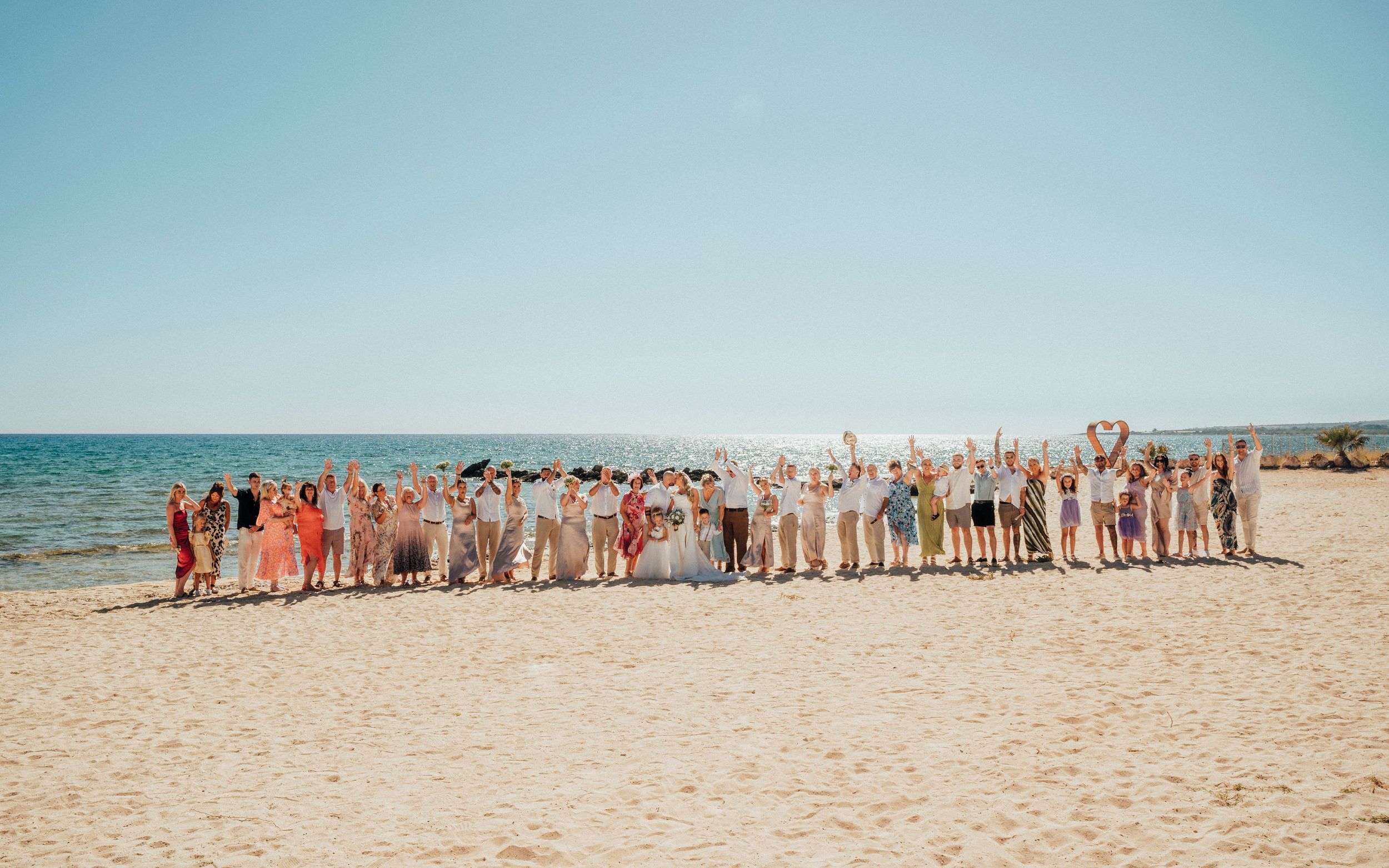 A large group of wedding guests poses together on a sunny beach with the ocean in the background.