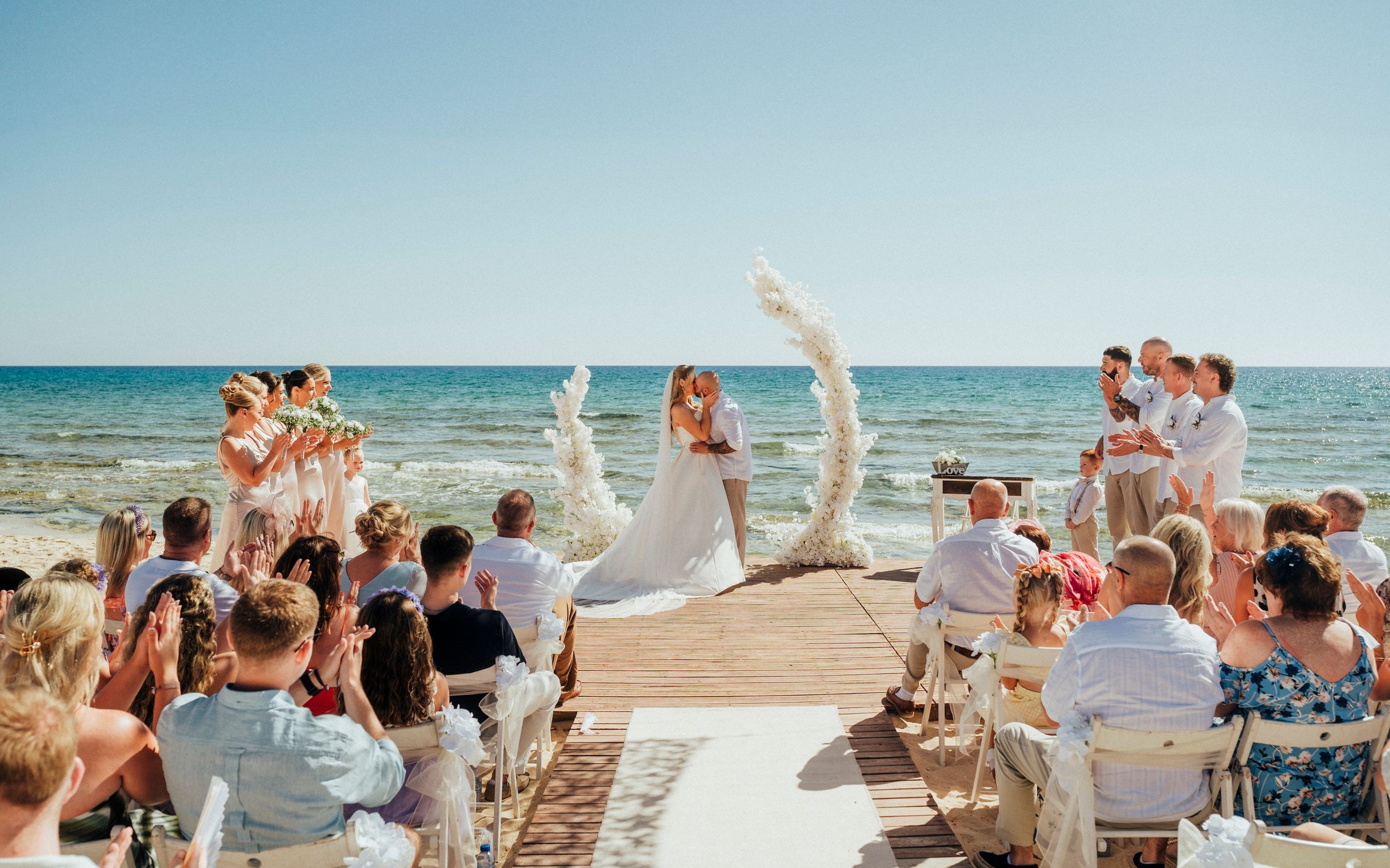 A joyful beach wedding ceremony unfolds on a wooden boardwalk as attendees in white celebrate under the blue summer sky.
