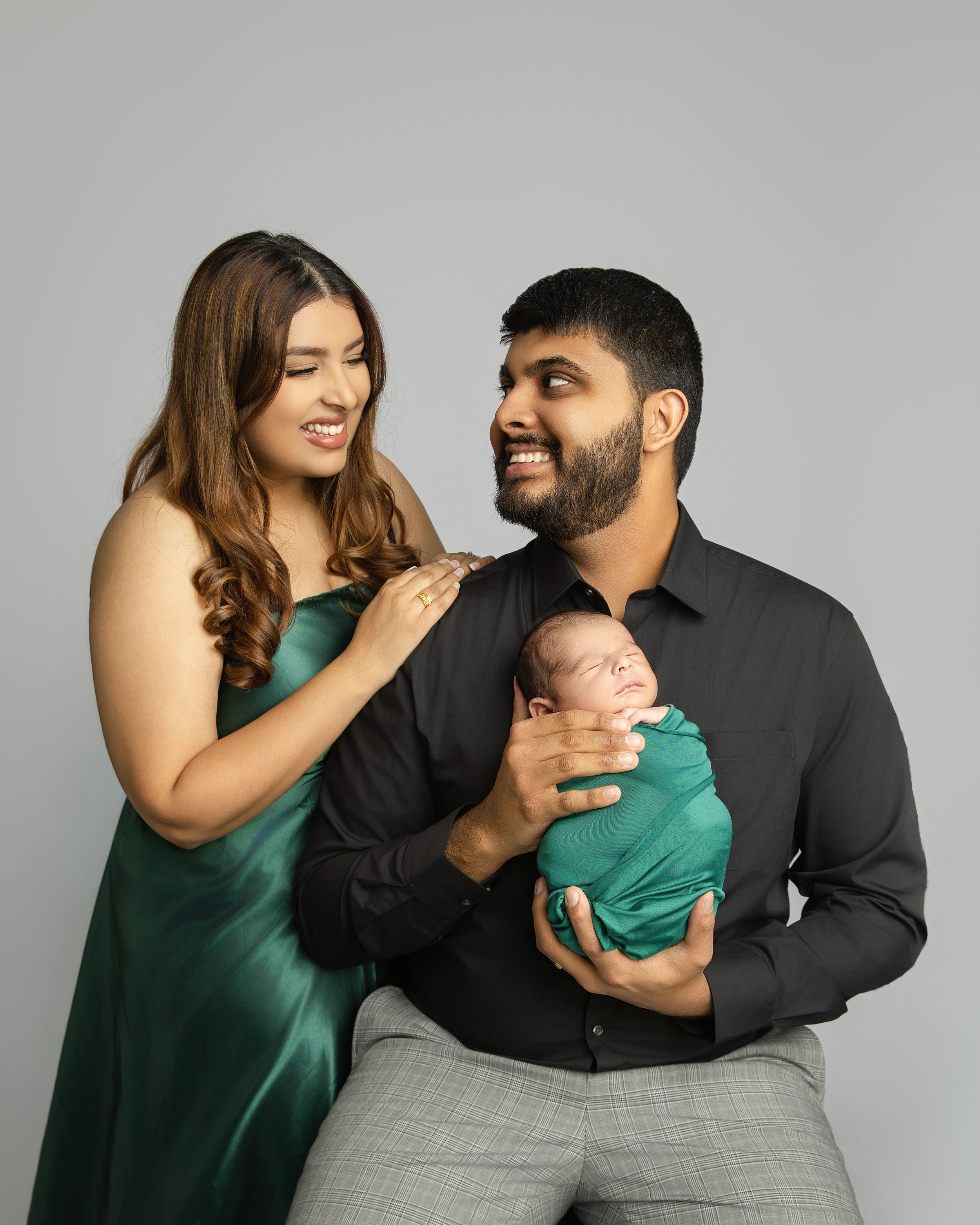 A couple in coordinating green and black outfits share tender moments while holding their newborn baby in a studio setting.
