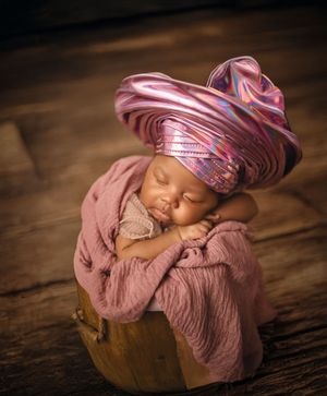 Sleeping newborn baby swaddled in mauve fabric lying in a wooden bowl against dark background.