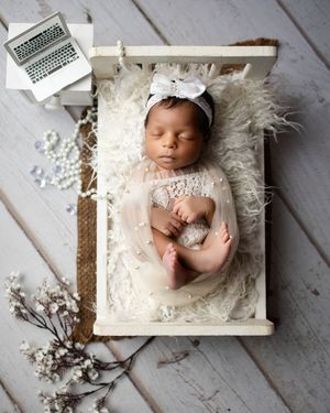 A sleeping newborn lies on white fluffy material in a rustic wooden crate with delicate floral accents and laptop props.