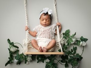 Newborn baby wearing white lace dress lies peacefully on wooden swing adorned with green and white floral decorations.