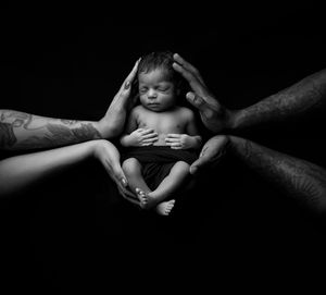 A black and white image of caring hands gently supporting a peaceful sleeping newborn baby against dark background.