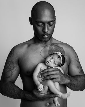 A tender black and white portrait of a shirtless person holding a sleeping baby in a gentle embrace.