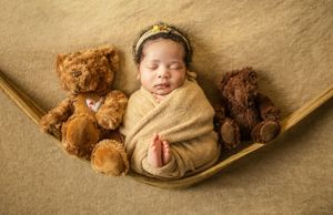 A sleeping baby in a hammock surrounded by plush teddy bears on a beige textured backdrop.