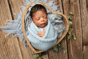 Newborn baby sleeping peacefully in blue knit wrap with wooden bowl prop on rustic background.