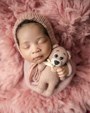 A sleeping newborn swaddled in pink blankets with a knitted teddy bear on a soft fluffy pink backdrop.