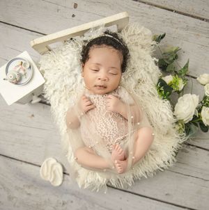 Sleeping baby in white lace dress lying on fluffy white rug with floral decorations on wooden floor.
