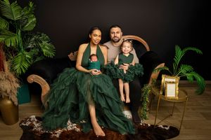 Family portrait with mother in green gown holding newborn, father, and young daughter in elegant studio setting.