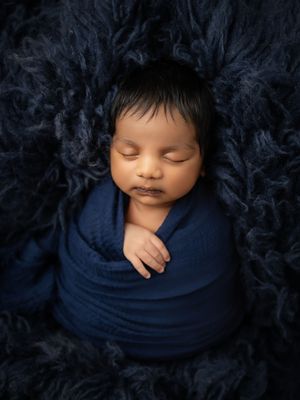 Peacefully sleeping newborn wrapped in navy blue swaddle blanket on dark fluffy fur background.
