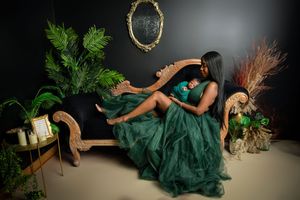 Elegant emerald green gown flows across vintage furniture amidst lush greenery and decor against a dark backdrop.