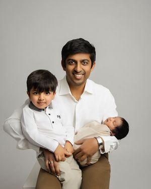 A family portrait against a gray background showing a father in white shirt holding a newborn while a toddler stands beside them.