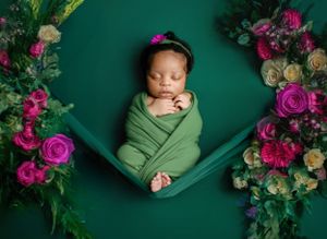 A newborn baby sleeps wrapped in green swaddle with vibrant pink and white flowers arranged on emerald backdrop.