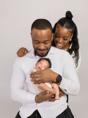 A couple in white outfits share a tender moment as they hold their sleeping newborn baby against a neutral backdrop.