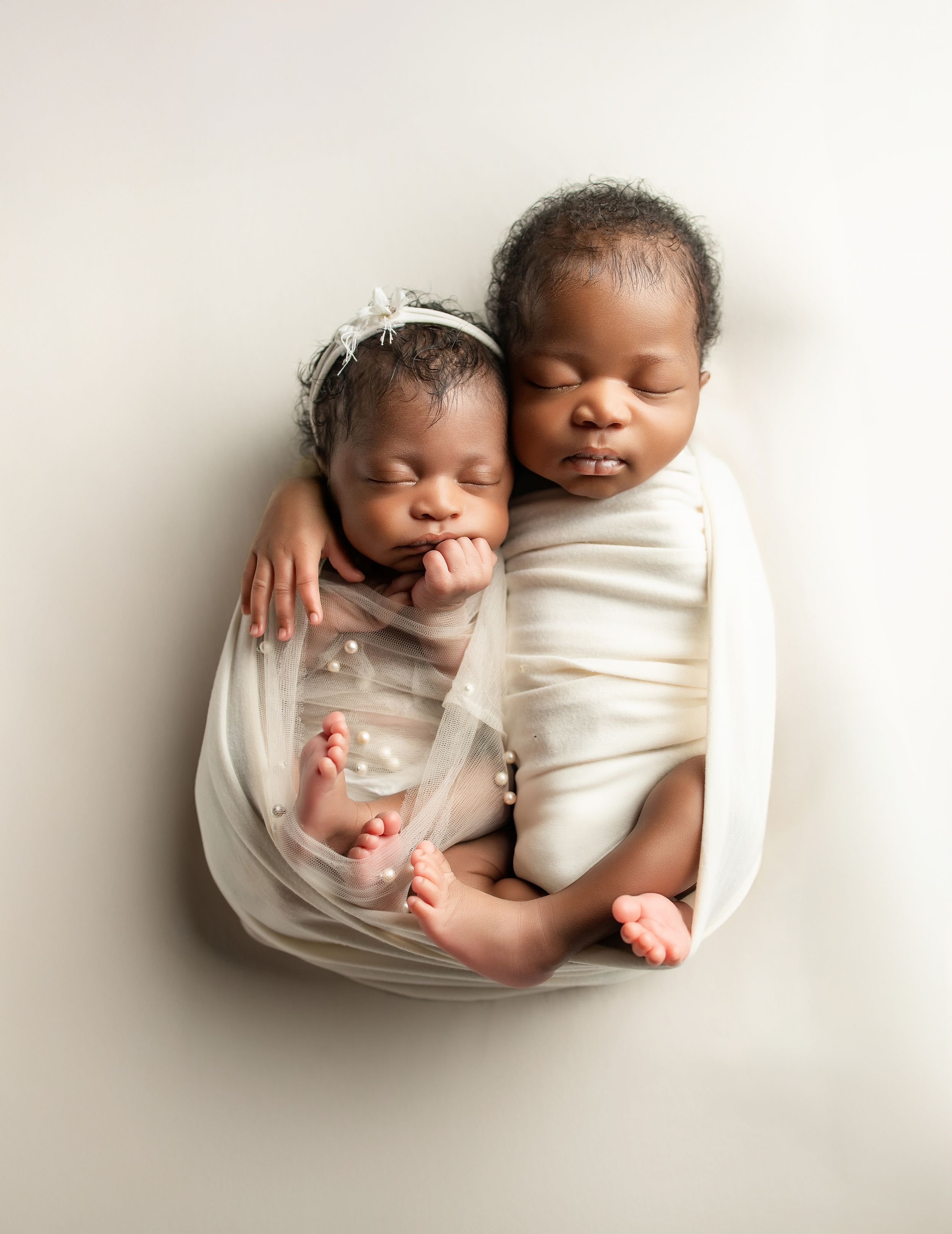 Two sleeping newborns swaddled in white wraps lie together peacefully on a neutral backdrop.