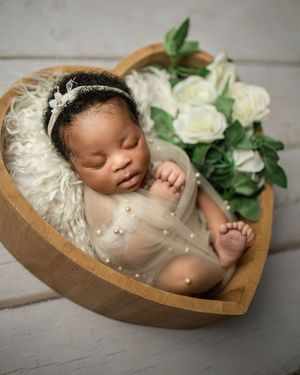 A sleeping infant reclines in a heart-shaped wooden bowl with white rose decorations beside them.