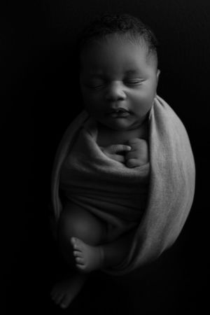Black and white artistic portrait of a sleeping newborn baby wrapped in soft fabric against dark background.