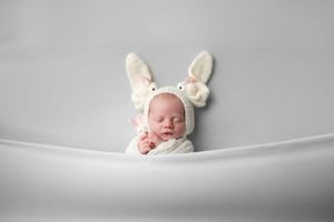 A sleeping baby wearing white bunny ears lies on a soft grey blanket in a minimalist newborn photography setup.
