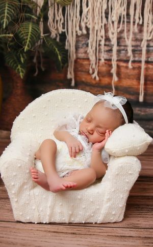 A sleeping baby dressed in white lace rests peacefully on a plush white armchair with macrame decor in the background.