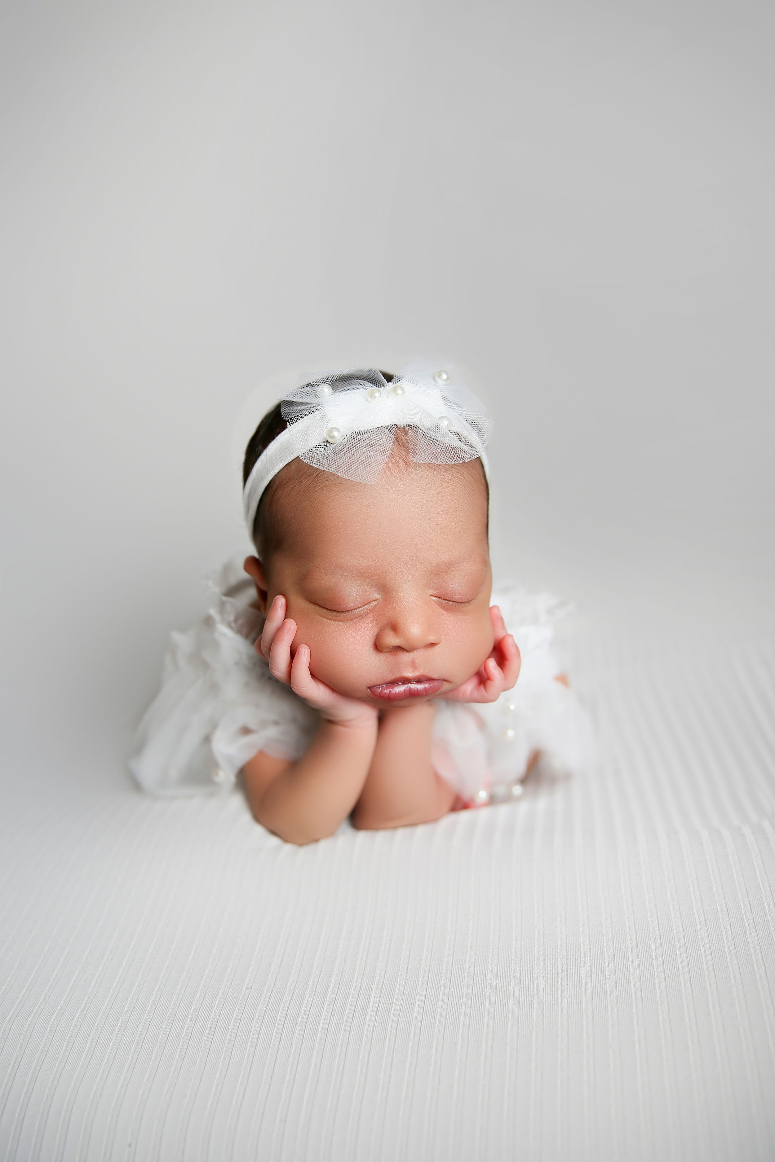 A sleeping newborn in white lace dress and headband rests head on hands in peaceful studio portrait.
