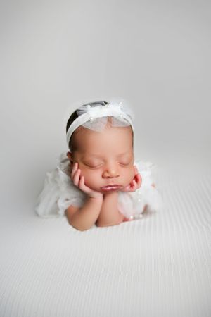 A sleeping newborn in white lace dress and headband rests head on hands in peaceful studio portrait.