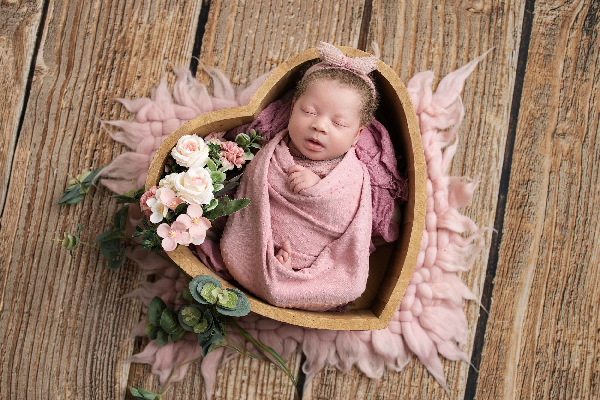 Newborn baby wrapped in pink swaddle sleeping in heart-shaped wooden bowl with flowers.