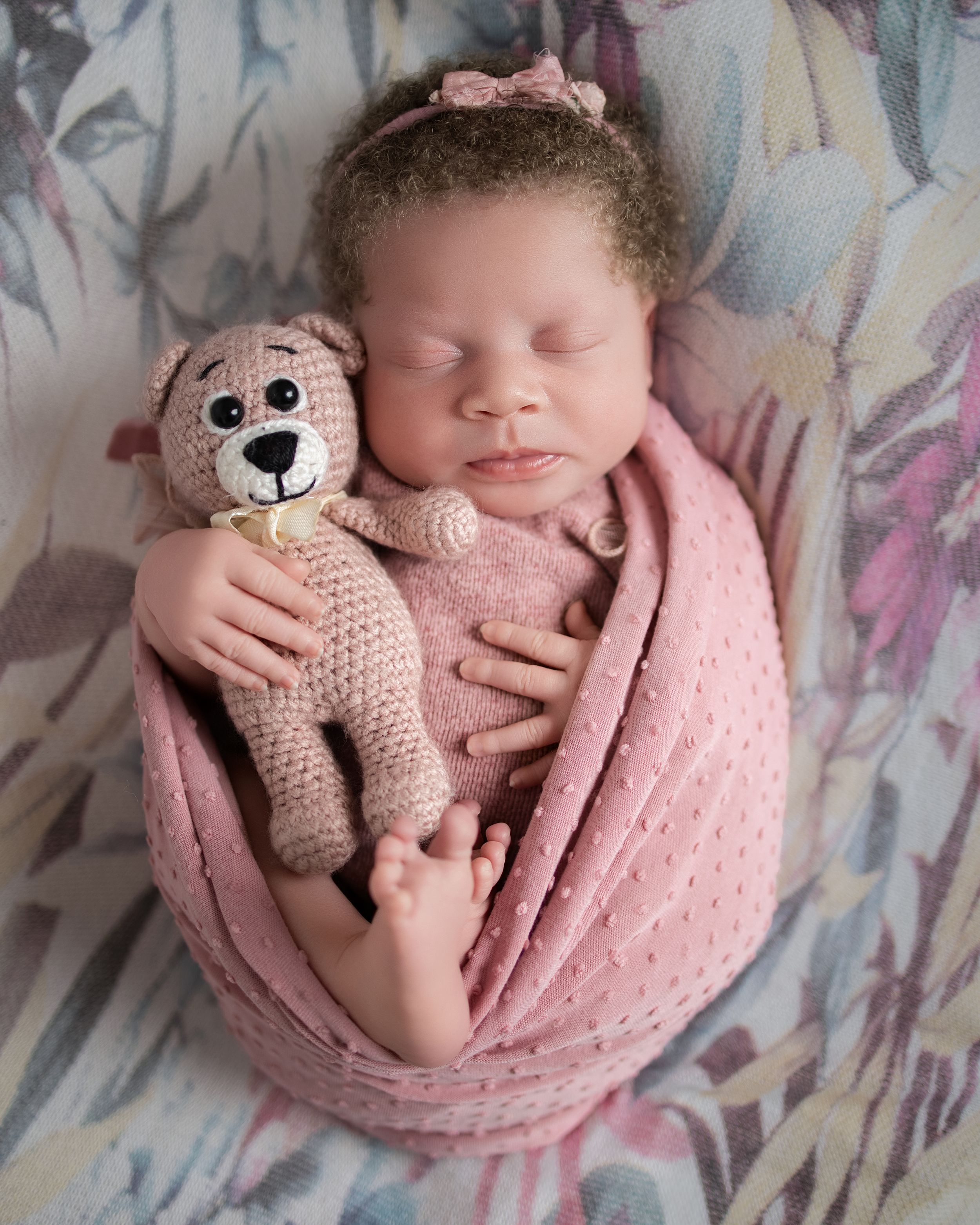 Newborn baby girl wrapped in pink blanket sleeping peacefully while holding a crocheted teddy bear with a pink bow headband.