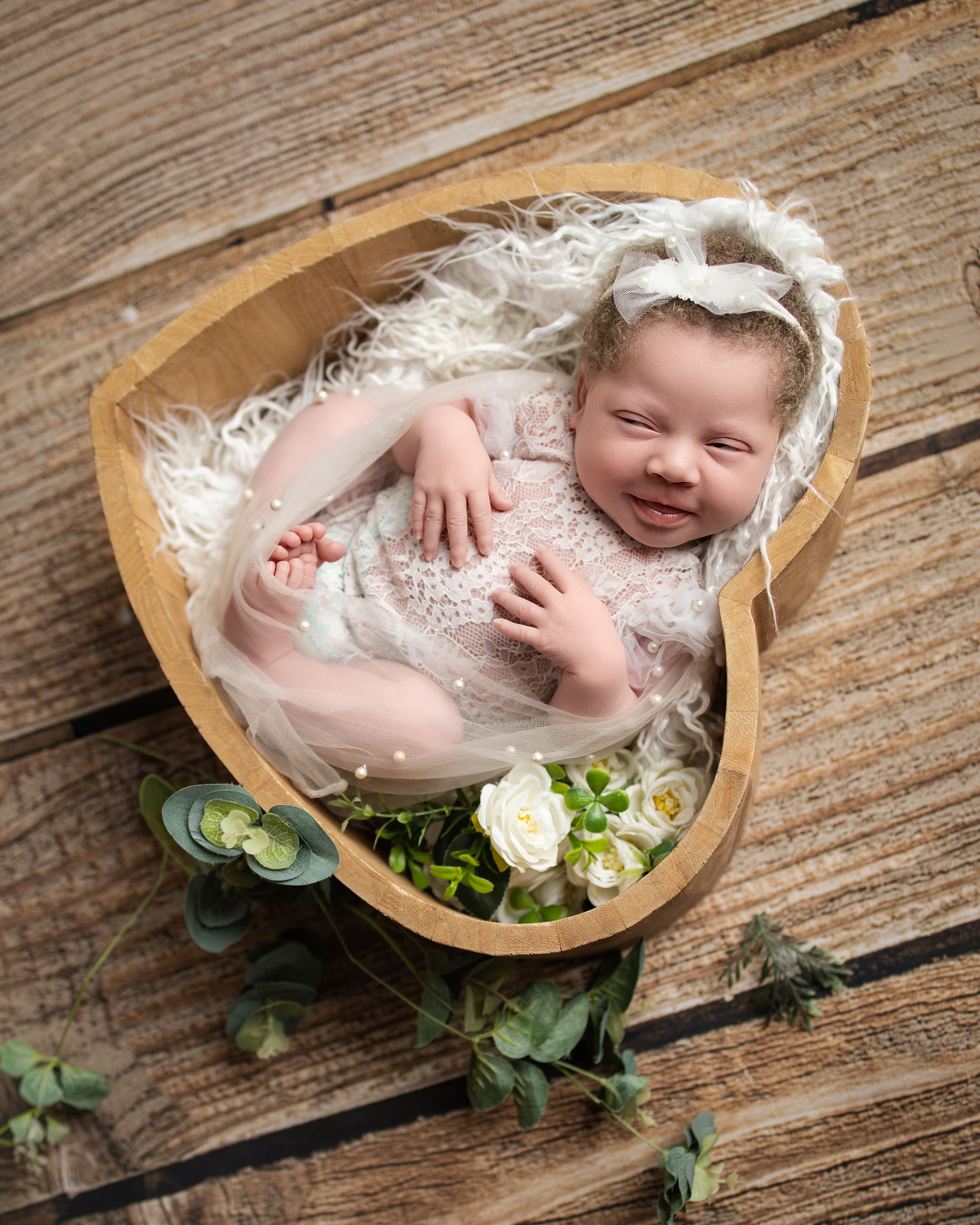 Smiling newborn baby in white lace outfit resting in a heart-shaped wooden bowl with flowers and greenery.