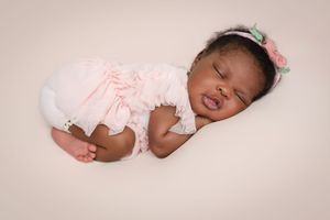 Sleeping newborn baby wearing soft white clothing lies peacefully on pink background in professional photo studio setting.