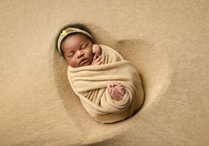 A sleeping newborn baby swaddled in beige fabric lies peacefully on a neutral backdrop during a professional photo session.