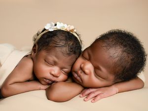 Two sleeping newborns in matching white outfits lay peacefully side by side on a beige backdrop.