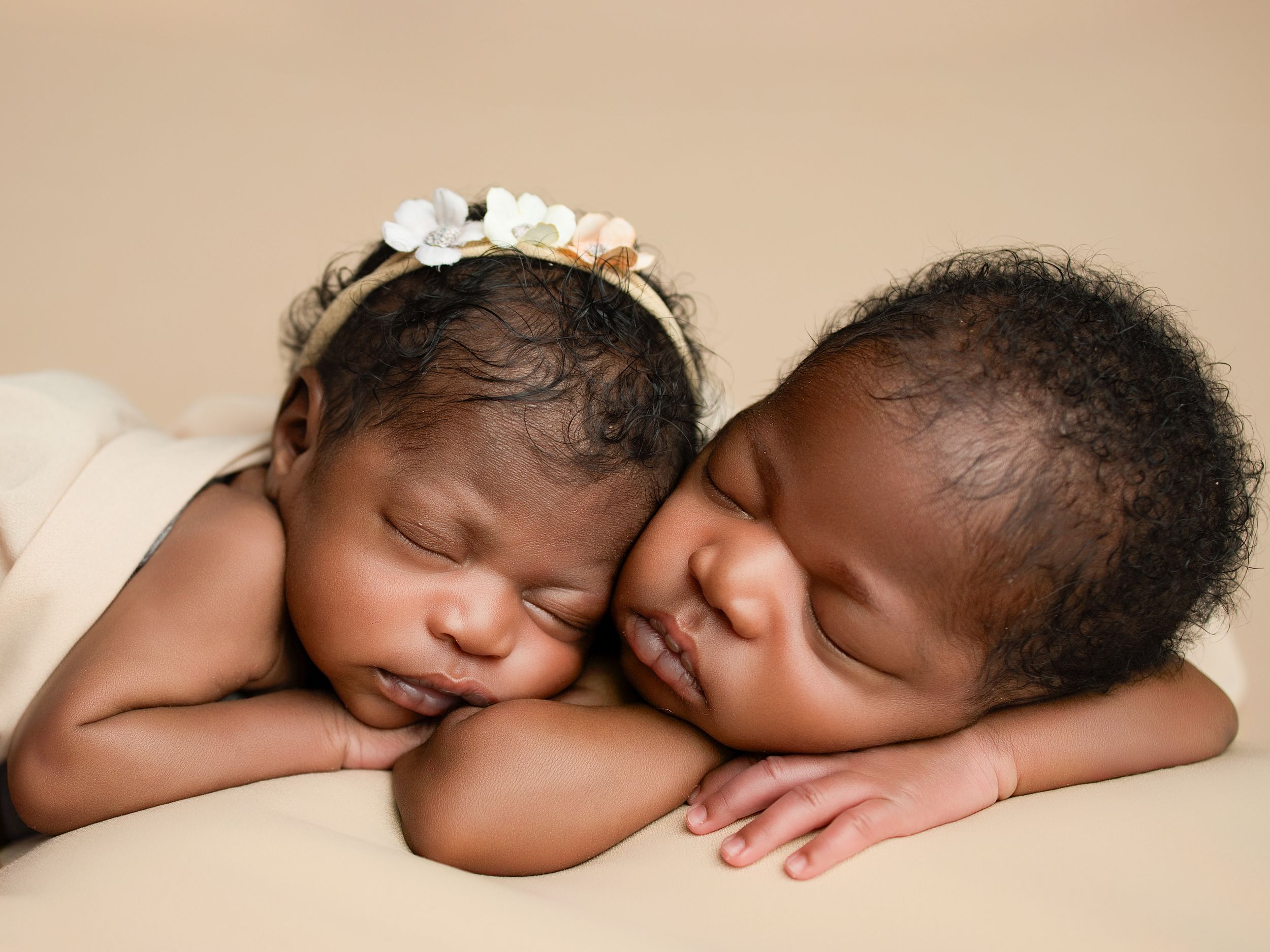 Two sleeping newborns in matching white outfits lay peacefully side by side on a beige backdrop.