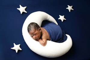 A sleeping newborn in a blue wrap rests peacefully on a white crescent moon prop with stars against a navy backdrop.