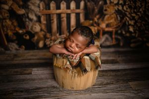 Sleeping baby curled up in brown wooden bucket with blanket against rustic wooden backdrop and picket fence.