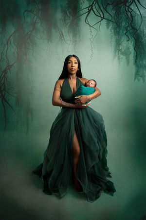 Elegant figure in flowing emerald green gown holds sleeping infant in mystical forest setting with moody lighting.