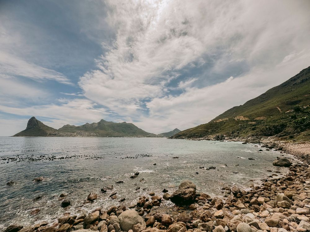 Rocky coastal landscape with turquoise waters and dramatic mountain range under a cloudy sky.