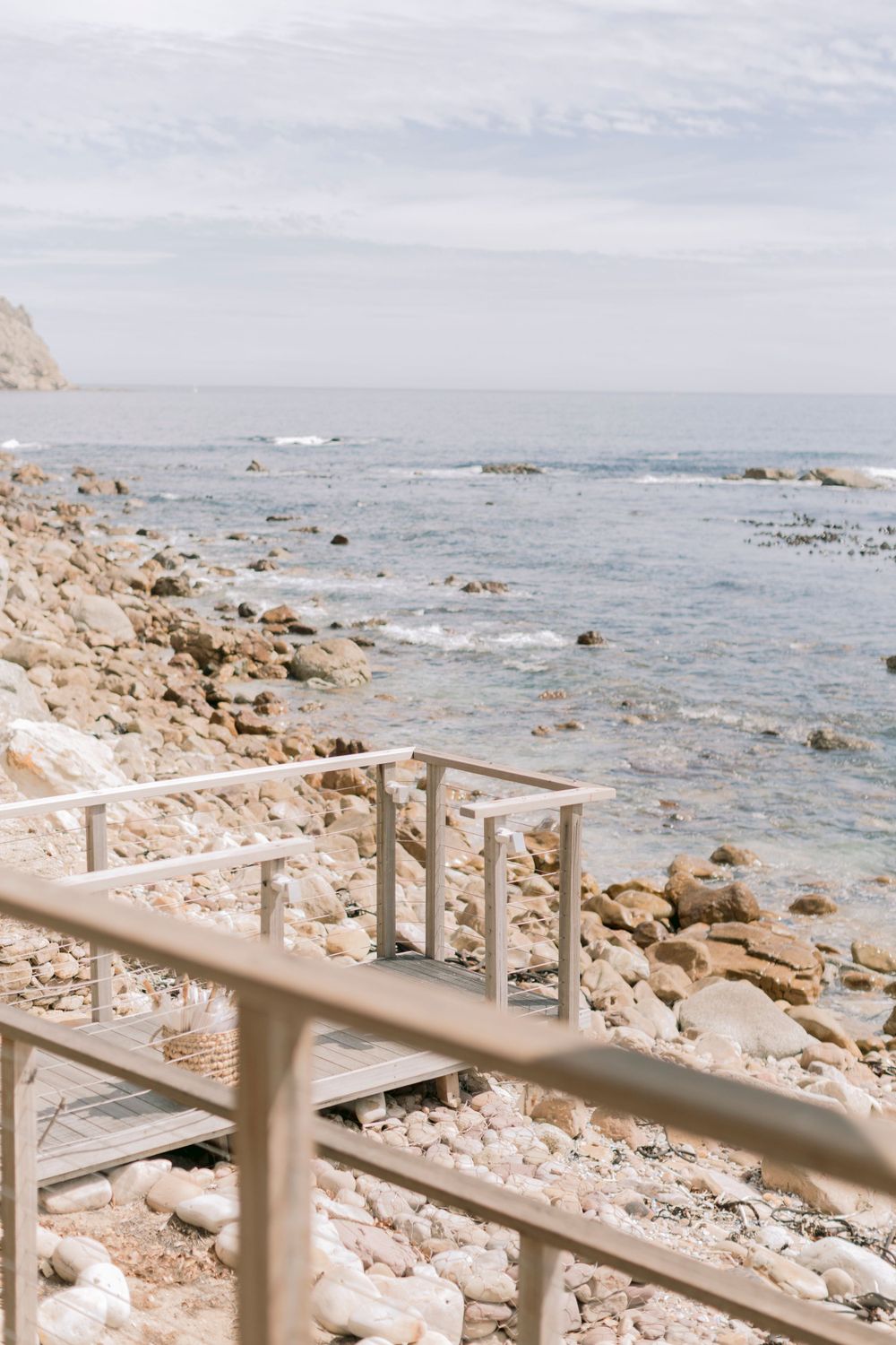 Wooden staircase with metal railings leading down to a rocky ocean shoreline with clear water.