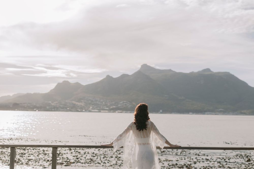 Woman in white dress stands at a railing gazing at a scenic mountain bay under soft overcast light.