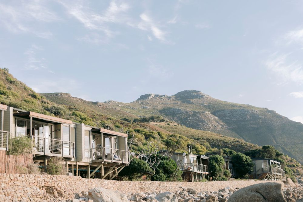 Modern beach cabins on stilts nestled against a rocky shore with a towering green mountain backdrop.