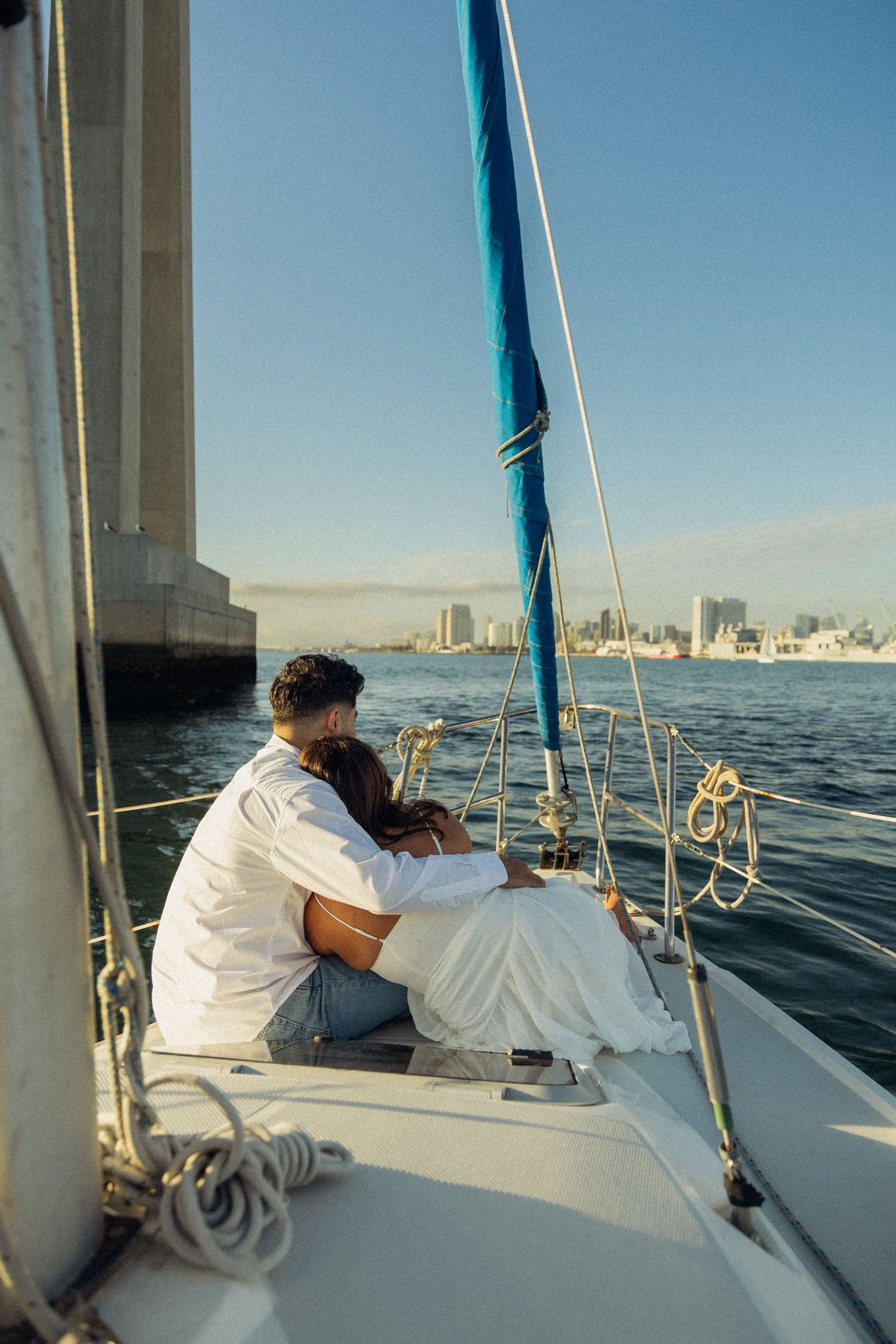 Sailboat passenger relaxes on deck while cruising past city skyline on calm waters during golden hour.