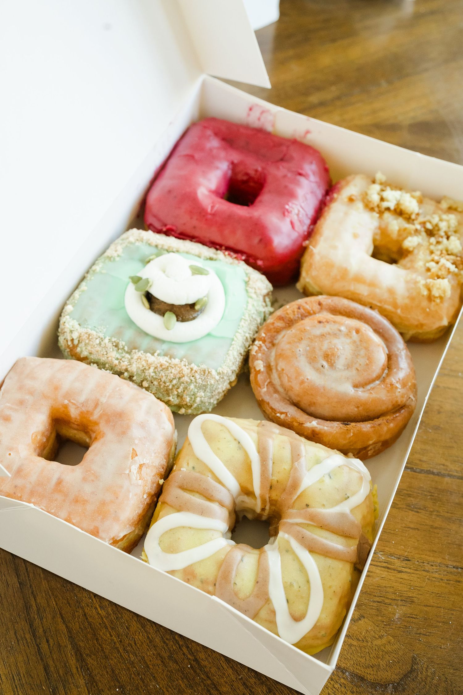 Six colorful artisan donuts with various toppings and glazes arranged neatly in a white bakery box.