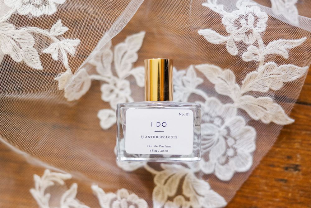 A glass perfume bottle with gold cap sits on a wooden surface with delicate white lace fabric.